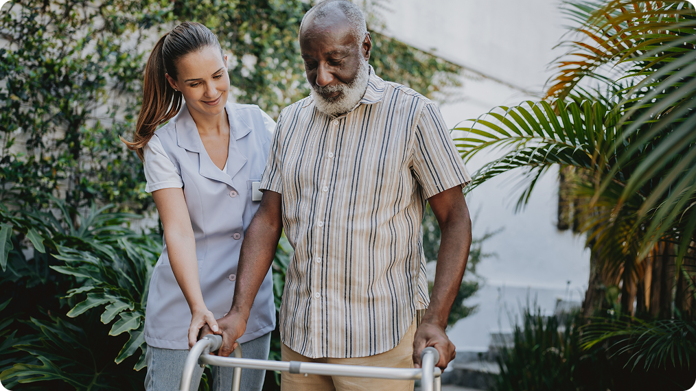 Therapist walking outdoors alongside an older man using a wheeled walker