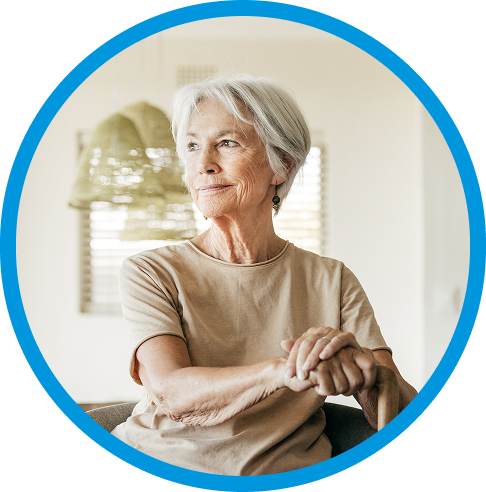 An older woman sitting at home, resting her hands on a walking cane and looking out a window